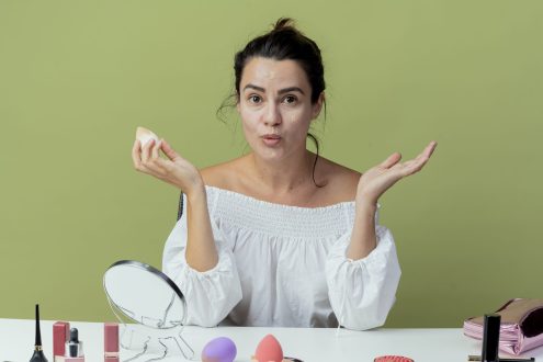 surprised beautiful girl sits at table with makeup tools applying foundation with sponge looking at camera isolated on green background with copy space